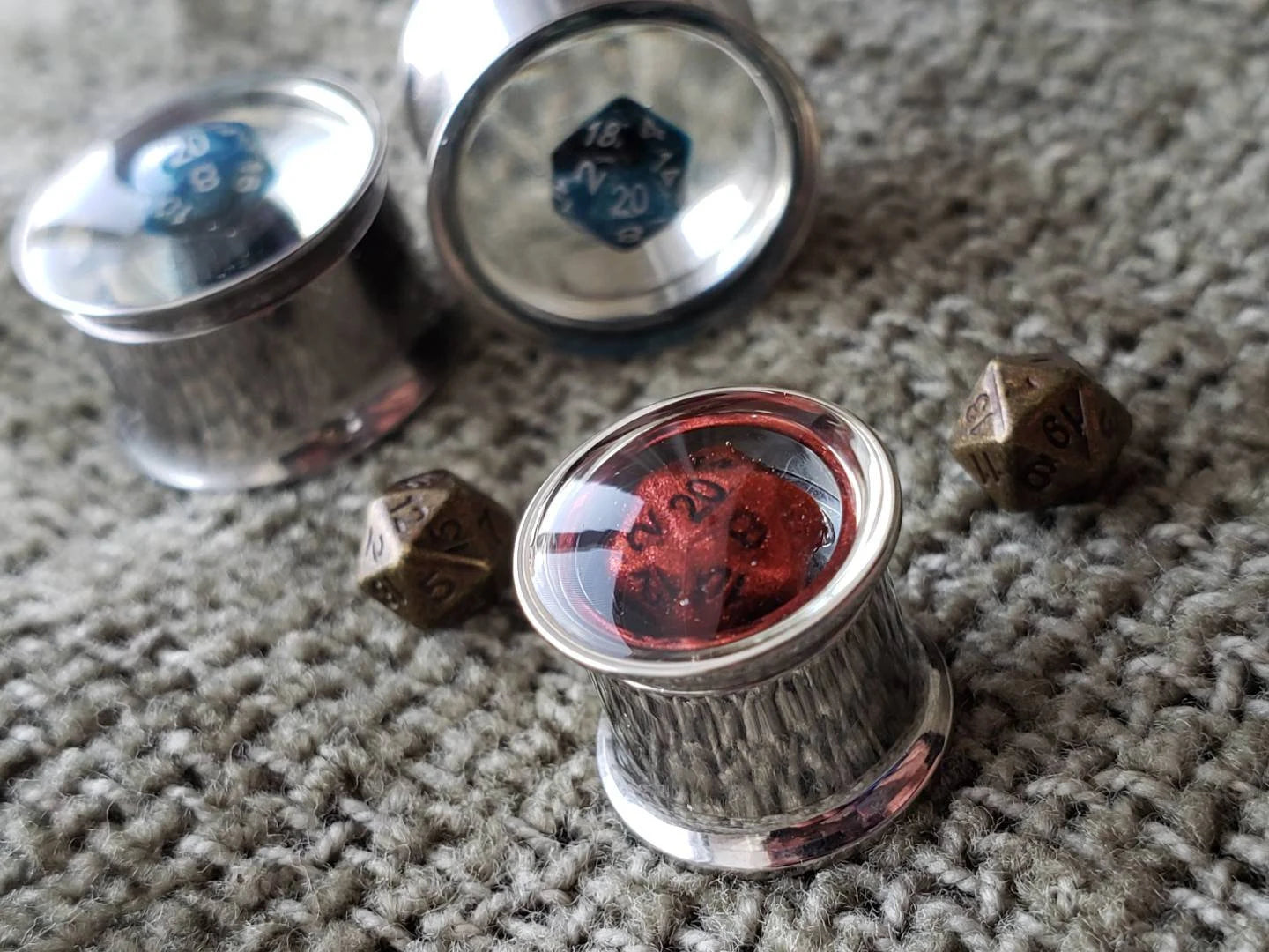 Stainless steel plugs for stretched ears, featuring a miniature blue D20 dice encased in clear resin. These gauges offer a sleek, modern look combined with a gaming-inspired design, ideal for tabletop RPG enthusiasts who love unique and high-quality jewelry. Photographed on a textured gray fabric background with a reflective metallic finish.