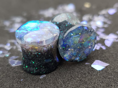 Close-up image of iridescent black, blue and green resin plugs for stretched ears, featuring sparkling flakes and a high-gloss finish. Perfect for body jewelry enthusiasts seeking unique and eye-catching ear gauges. Displayed on a black textured background with scattered glitter accents.