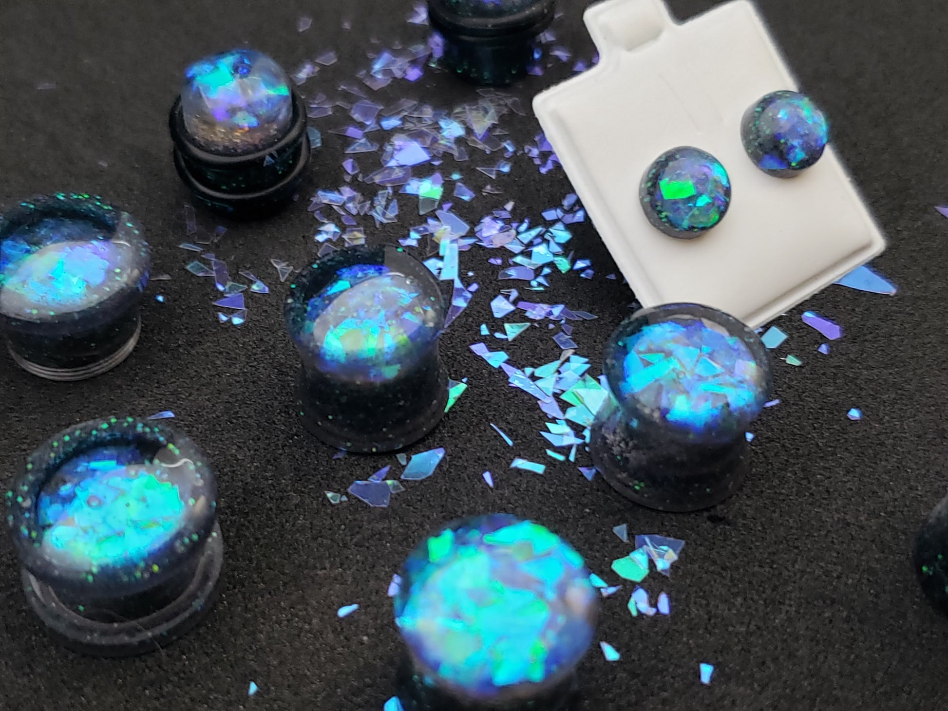 Close-up image of iridescent black, blue and green resin plugs for stretched ears, featuring sparkling flakes and a high-gloss finish. Perfect for body jewelry enthusiasts seeking unique and eye-catching ear gauges. Displayed on a black textured background with scattered glitter accents.