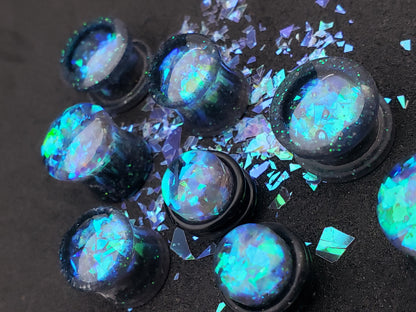 Close-up image of iridescent black, blue and green resin plugs for stretched ears, featuring sparkling flakes and a high-gloss finish. Perfect for body jewelry enthusiasts seeking unique and eye-catching ear gauges. Displayed on a black textured background with scattered glitter accents.