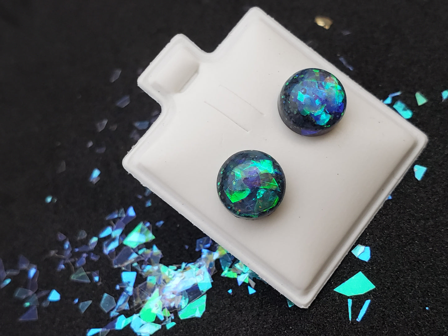 Close-up image of iridescent black, blue and green resin earrings for pierced ears, featuring sparkling flakes and a high-gloss finish. Perfect for body jewelry enthusiasts seeking unique and eye-catching earrings. Displayed on a black textured background with scattered glitter accents.