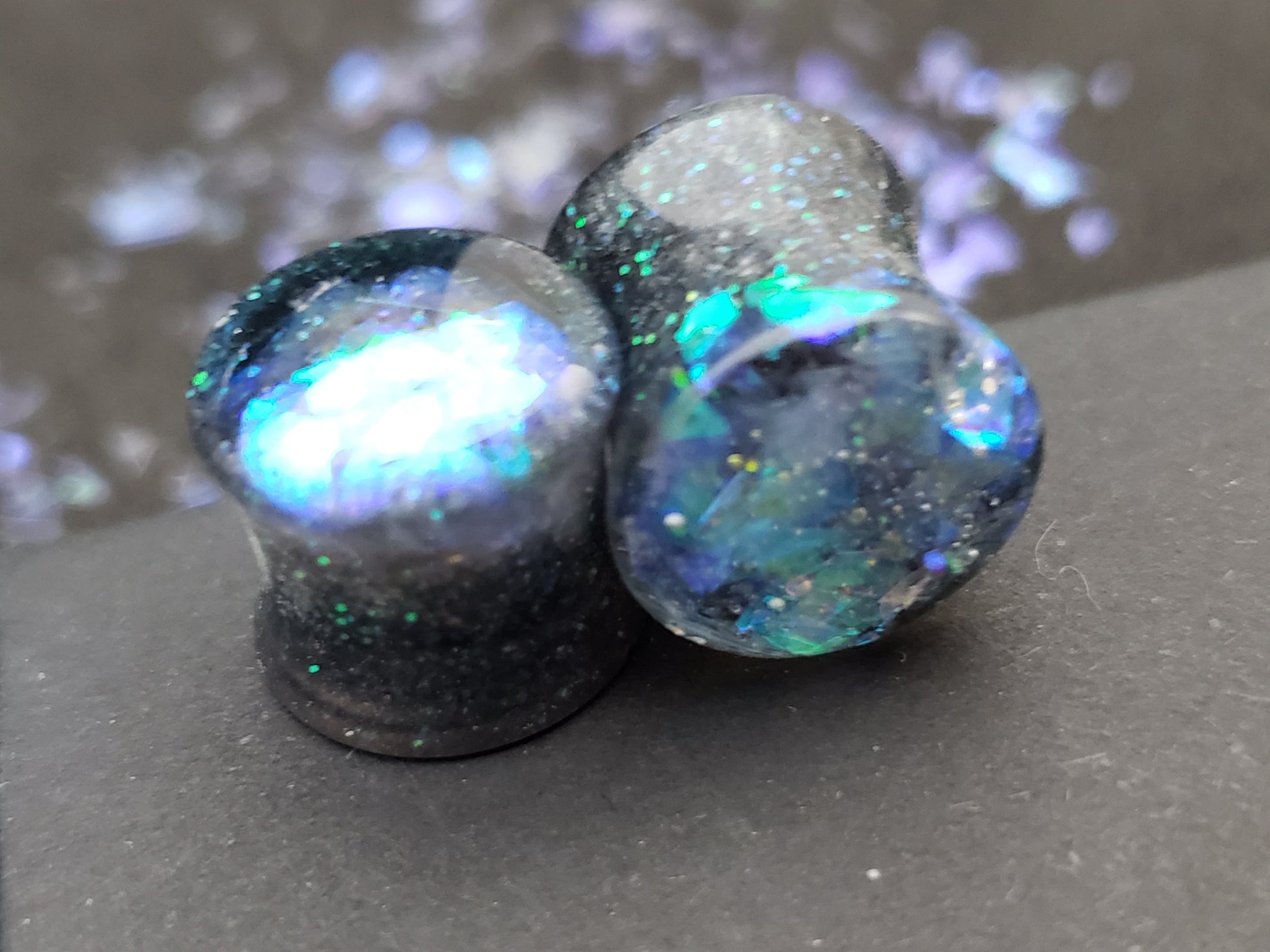 Close-up image of iridescent black, blue and green resin plugs for stretched ears, featuring sparkling flakes and a high-gloss finish. Perfect for body jewelry enthusiasts seeking unique and eye-catching ear gauges. Displayed on a black textured background with scattered glitter accents.