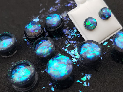 Close-up image of iridescent black, blue and green resin plugs for stretched ears, featuring sparkling flakes and a high-gloss finish. Perfect for body jewelry enthusiasts seeking unique and eye-catching ear gauges. Displayed on a black textured background with scattered glitter accents.