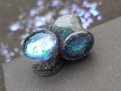 Close-up image of iridescent black, blue and green resin plugs for stretched ears, featuring sparkling flakes and a high-gloss finish. Perfect for body jewelry enthusiasts seeking unique and eye-catching ear gauges. Displayed on a black textured background with scattered glitter accents.