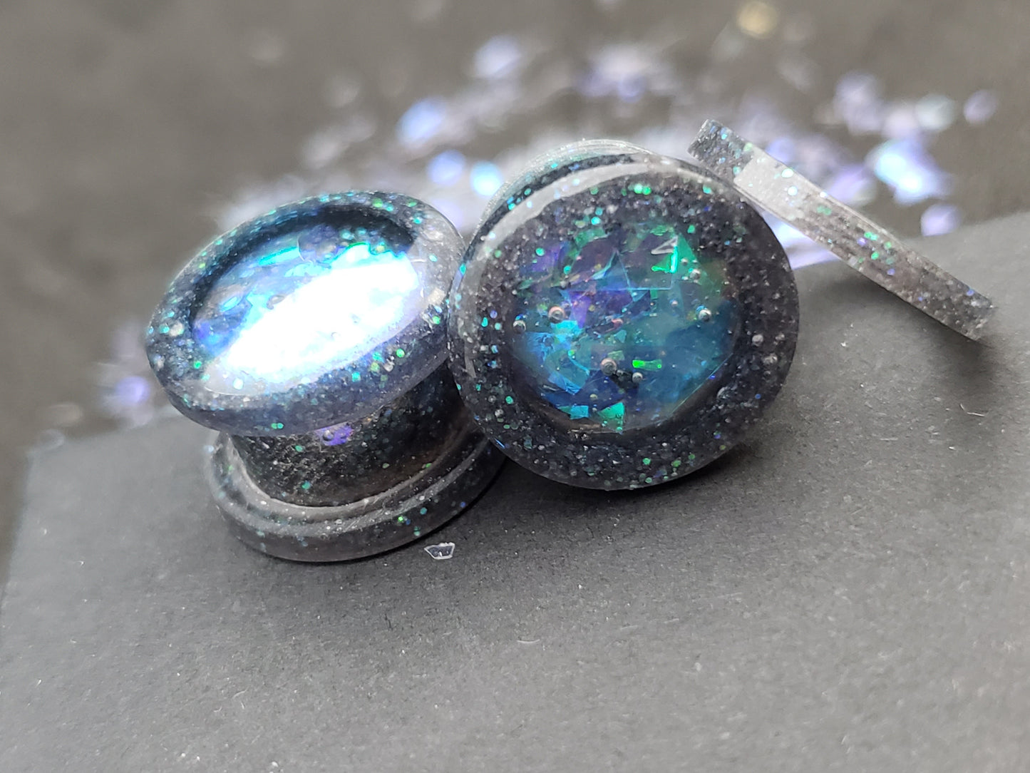 Close-up image of iridescent black, blue and green resin plugs for stretched ears, featuring sparkling flakes and a high-gloss finish. Perfect for body jewelry enthusiasts seeking unique and eye-catching ear gauges. Displayed on a black textured background with scattered glitter accents.