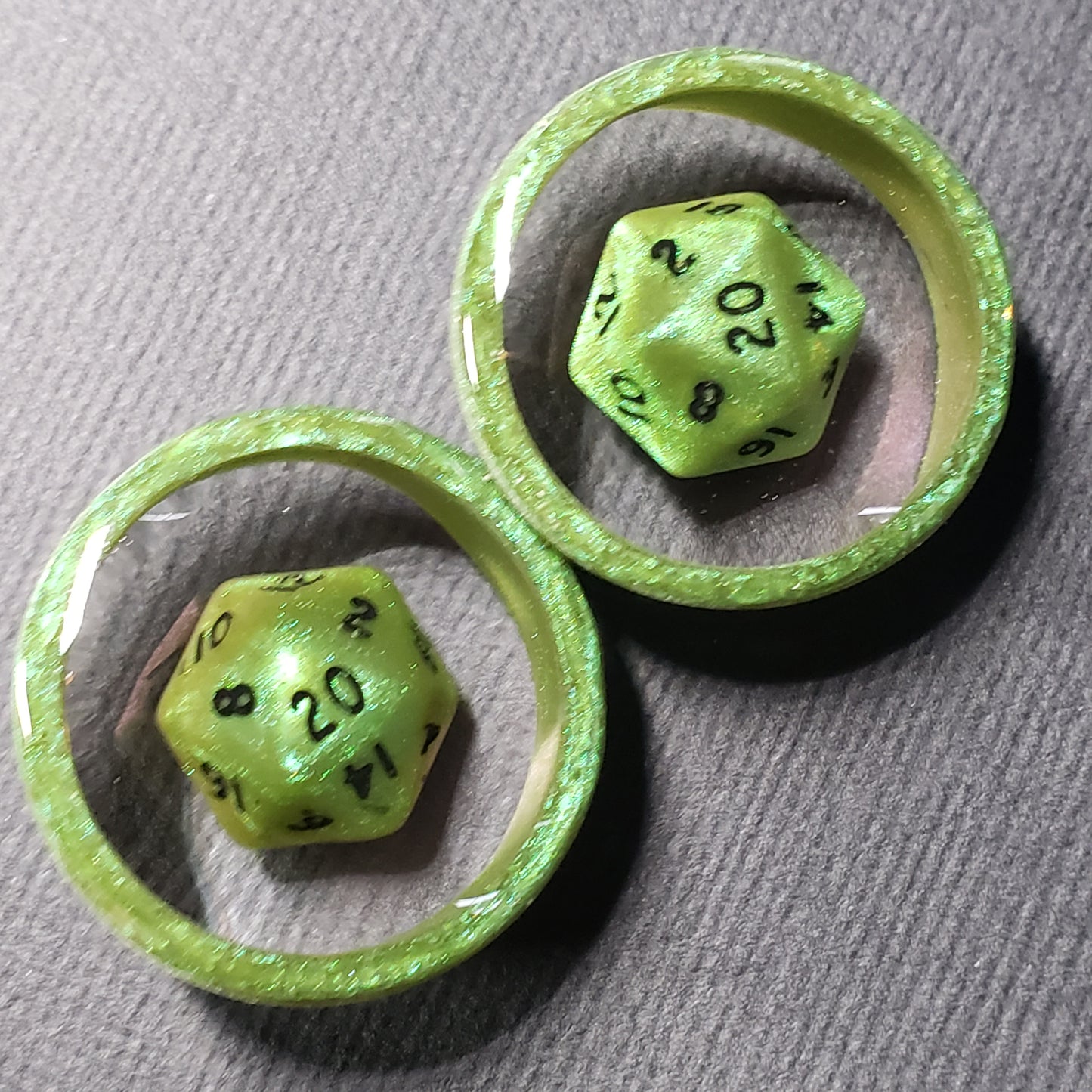 Green resin plugs for stretched ears, featuring a miniature D20 dice encased in clear resin. These handcrafted gauges combine a playful tabletop gaming theme with a subtle, elegant pink shimmer. Ideal for RPG fans and unique jewelry collectors, photographed on a soft, textured gray fabric background.