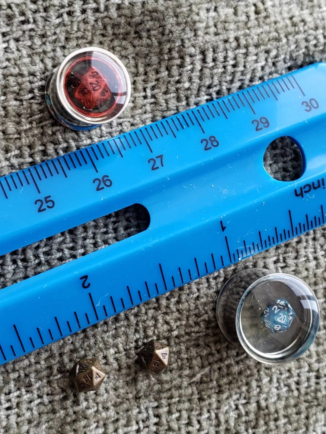 Stainless steel plugs for stretched ears, featuring a miniature blue D20 dice encased in clear resin. These gauges offer a sleek, modern look combined with a gaming-inspired design, ideal for tabletop RPG enthusiasts who love unique and high-quality jewelry. Photographed on a textured gray fabric background with a reflective metallic finish.