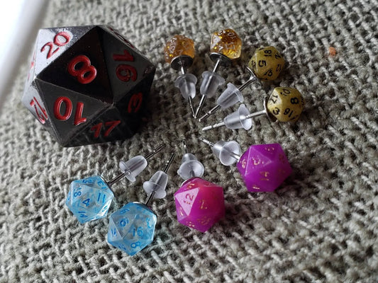 Set of colorful D20 dice earrings featuring mini dice in blue, pink, gold, and amber tones, with stainless steel posts and silicone backings. A large black and red D20 dice accentuates the gaming theme, making these earrings a perfect accessory for tabletop RPG fans. Photographed on a soft gray fabric background to highlight their vibrant colors and intricate details.