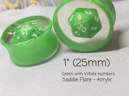 Green resin plugs for stretched ears, featuring a miniature D20 dice encased in clear resin. These handcrafted gauges combine a playful tabletop gaming theme with a subtle, elegant pink shimmer. Ideal for RPG fans and unique jewelry collectors, photographed on a soft, textured gray fabric background.