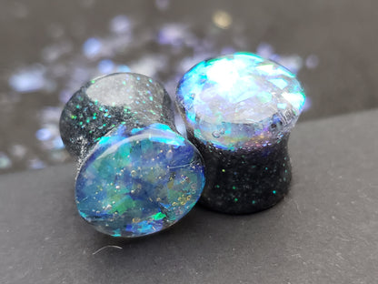 Close-up image of iridescent black, blue and green resin plugs for stretched ears, featuring sparkling flakes and a high-gloss finish. Perfect for body jewelry enthusiasts seeking unique and eye-catching ear gauges. Displayed on a black textured background with scattered glitter accents.