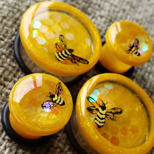 Yellow resin plugs for stretched ears, featuring a detailed bee design over a honeycomb-inspired pattern with holographic accents. These handcrafted gauges are perfect for nature enthusiasts and alternative jewelry lovers. Captured on a textured fabric background, highlighting their vibrant and whimsical design.