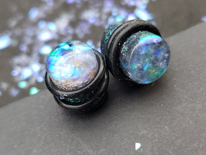 Close-up image of no flare iridescent black, blue and green resin plugs for stretched ears, featuring sparkling flakes and a high-gloss finish. Perfect for body jewelry enthusiasts seeking unique and eye-catching ear gauges. Displayed on a black textured background with scattered glitter accents.