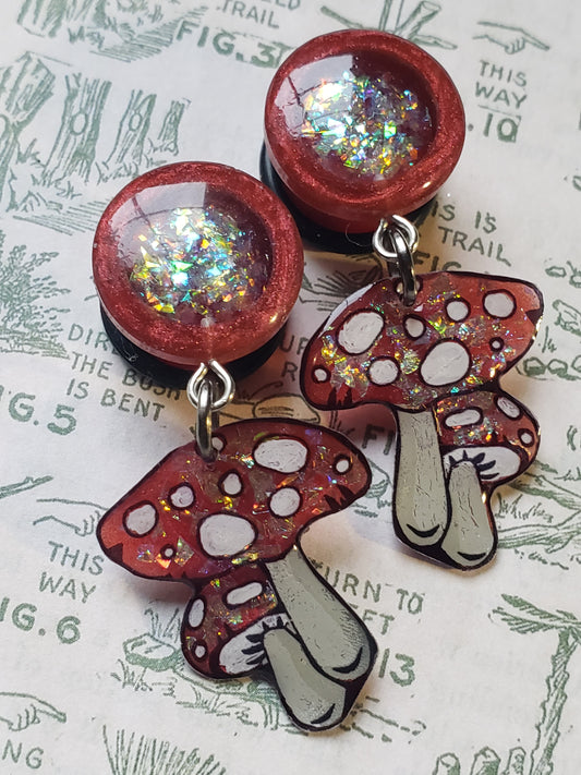 Handmade red resin dangle plugs with glittery mushroom-shaped charms for stretched ears. The plugs are accented with holographic flakes, making them ideal for alternative jewelry lovers who enjoy whimsical and nature-inspired designs. Set against a floral-themed backdrop for a stylish aesthetic.