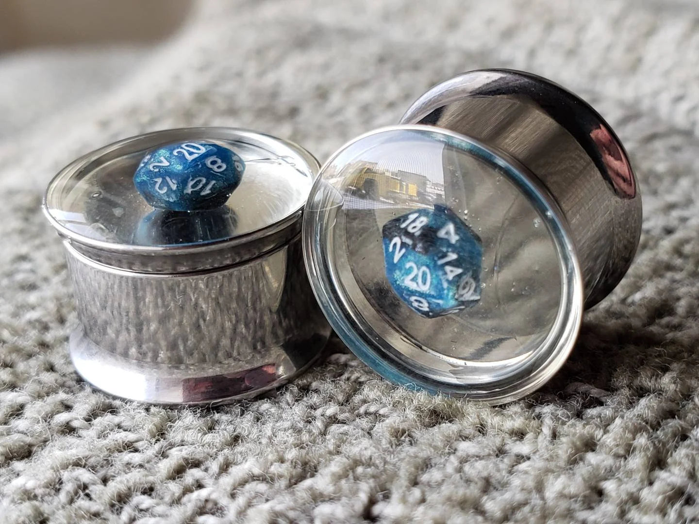 Stainless steel plugs for stretched ears, featuring a miniature blue D20 dice encased in clear resin. These gauges offer a sleek, modern look combined with a gaming-inspired design, ideal for tabletop RPG enthusiasts who love unique and high-quality jewelry. Photographed on a textured gray fabric background with a reflective metallic finish.